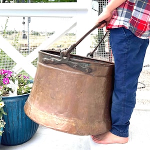 May include: A large, round, antique copper bucket with a wide opening and a tapered base. The bucket features a dark, weathered metal handle attached to the rim with riveted brackets. The copper surface shows a natural, aged patina with visible texture.