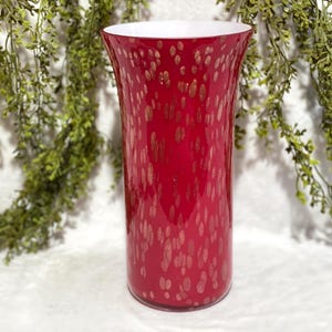 Puede incluir: Un jarrón alto de cristal rojo con interior blanco y motas doradas. El jarrón tiene una parte superior acampanada y un cuerpo cilíndrico. Hay vegetación decorativa en el fondo.