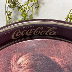 Może przedstawiać: Zabytkowy taca do serwowania Coca-Cola z bordowym obramowaniem i złotymi literami. Taca zawiera logo Coca-Cola i napis "DRINK Coca-Cola". Taca ma wyblakły obrazek pośrodku.