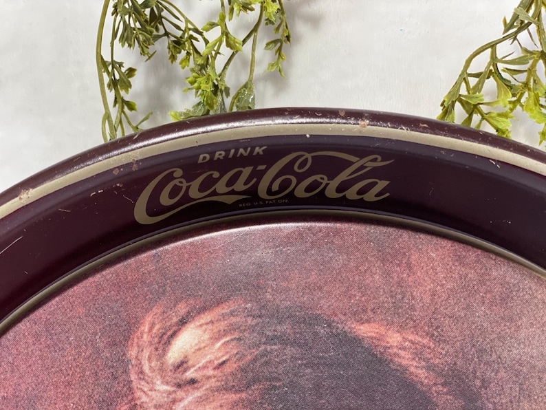 Może przedstawiać: Zabytkowy taca do serwowania Coca-Cola z bordowym obramowaniem i złotymi literami. Taca zawiera logo Coca-Cola i napis "DRINK Coca-Cola". Taca ma wyblakły obrazek pośrodku.