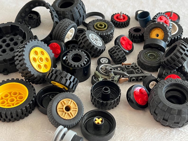 May include: A collection of black toy wheels of various sizes, some with yellow, red, or gold centers. The tires have textured treads. There are also other toy parts in the image.