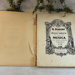May include: An open vintage music book with a cream-colored cover and pages. The cover features ornate black text and illustrations, including the title "R. Dimuro Metodo Completo della Musica". The book is slightly worn, with visible signs of age.