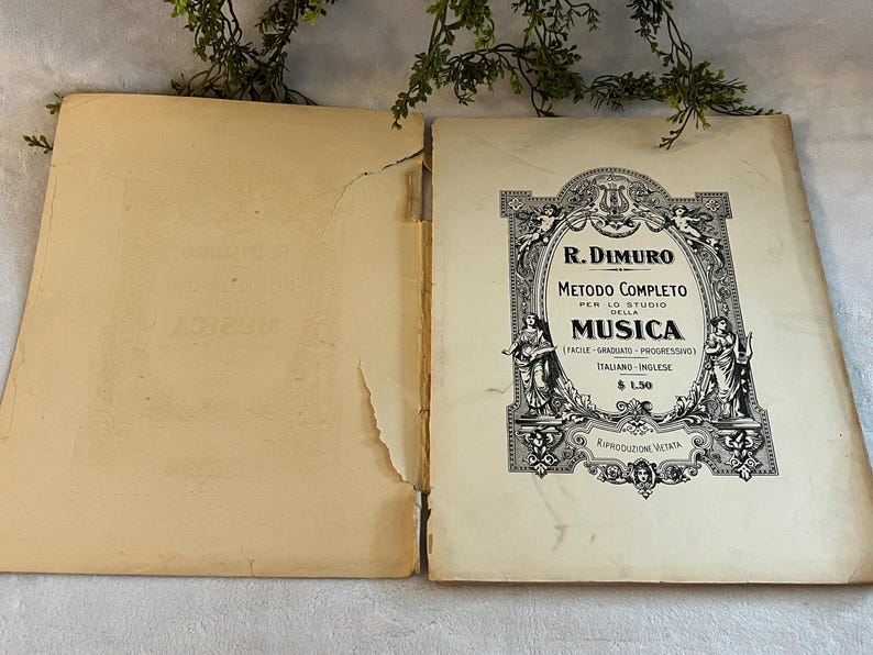May include: An open vintage music book with a cream-colored cover and pages. The cover features ornate black text and illustrations, including the title "R. Dimuro Metodo Completo della Musica". The book is slightly worn, with visible signs of age.