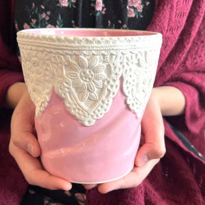 May include: A pink ceramic planter with a white lace design around the top. The planter is held in both hands. The lace design features floral patterns. The interior of the planter is also pink.