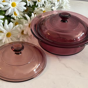 May include: A round, translucent purple glass casserole dish with a matching lid and a separate, identical glass lid. Both lids feature a central knob and a radiating sunburst pattern, while the dish body has vertical ribbed detailing.
