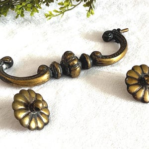 May include: Antique brass-toned drawer pulls and knobs. The set includes a long, curved handle with ornate detailing and two matching flower-shaped knobs. The hardware has a vintage aesthetic, with a warm, aged finish.