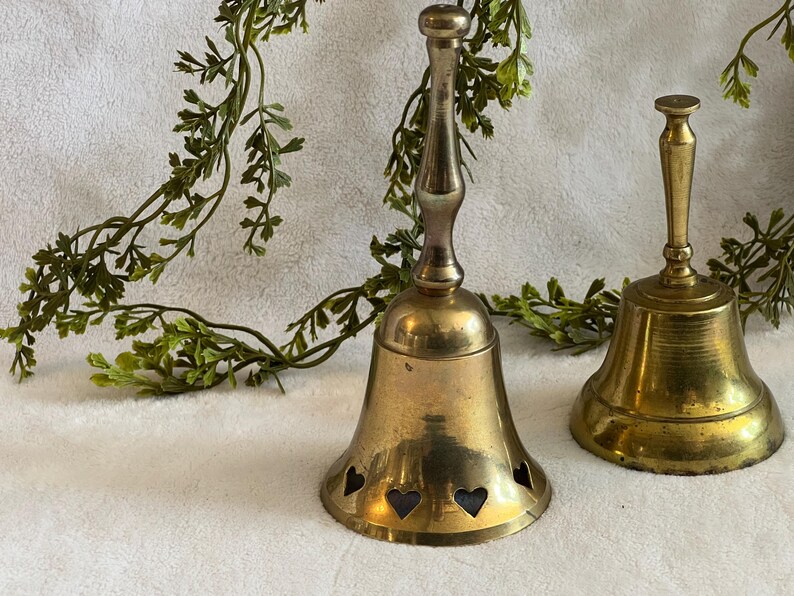May include: Two antique brass bells. The larger bell has heart-shaped cutouts near the base. Both bells have a tarnished, aged appearance. Greenery is draped in the background, adding a touch of nature to the scene.