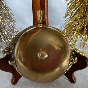 Op de afbeelding: Een messing bel met een decoratief ontwerp rond de rand, gemonteerd op een houten standaard. De bel heeft een glad, glanzend oppervlak en een kleine knop aan de bovenkant.
