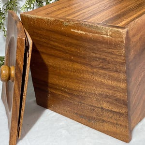 May include: A rectangular wooden box with a lid, showcasing a warm brown hue and visible wood grain. The lid, with a rounded knob, is slightly ajar, revealing the interior. The box appears to be vintage, with some wear and tear visible.