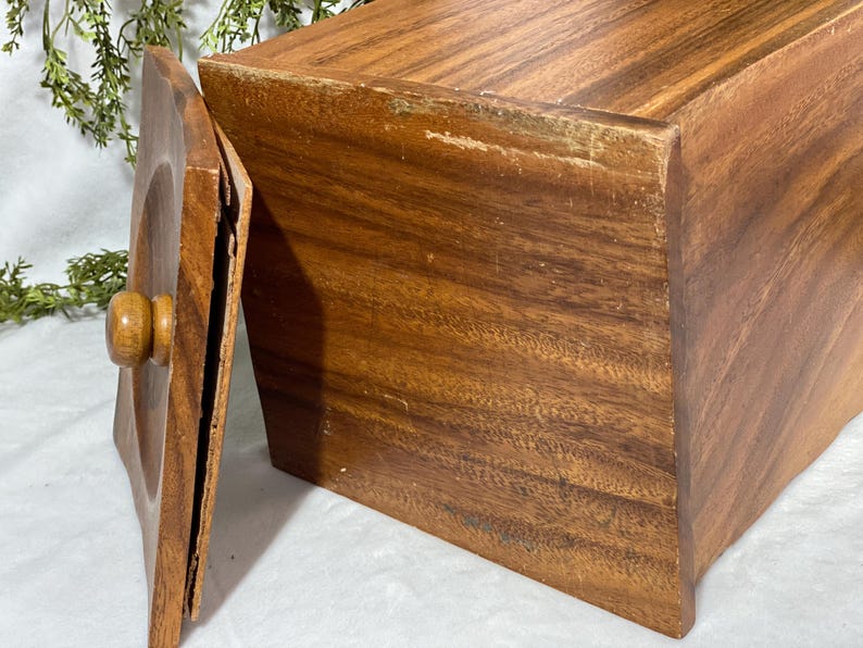 May include: A rectangular wooden box with a lid, showcasing a warm brown hue and visible wood grain. The lid, with a rounded knob, is slightly ajar, revealing the interior. The box appears to be vintage, with some wear and tear visible.