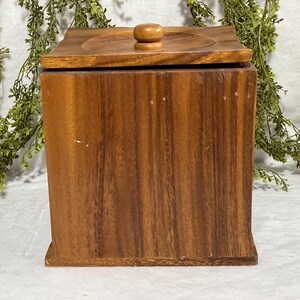May include: A square wooden box with a lid. The box is made of a dark brown wood with a visible grain pattern. The lid has a circular indentation and a small, round wooden knob. The box is likely used for storage.