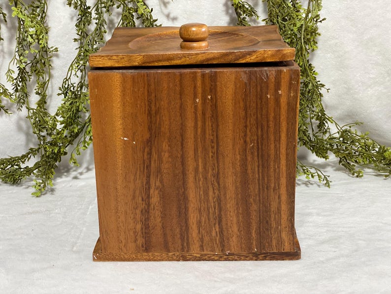 May include: A square wooden box with a lid. The box is made of a dark brown wood with a visible grain pattern. The lid has a circular indentation and a small, round wooden knob. The box is likely used for storage.