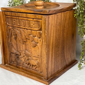 May include: A wooden flour container with a lid and a carved landscape design. The word "FLOUR" is carved on the front. The container is made of a warm brown wood and has a square shape.