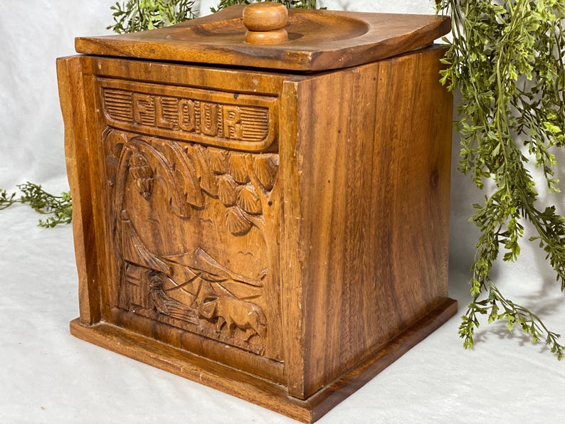 May include: A wooden flour container with a lid and a carved landscape design. The word "FLOUR" is carved on the front. The container is made of a warm brown wood and has a square shape.