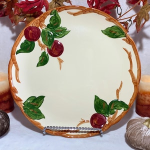 Puede incluir: Plato decorativo color crema con un borde marrón en forma de rama. El plato presenta manzanas rojas y hojas verdes. El plato se exhibe sobre un soporte plateado, con calabazas y velas en el fondo.