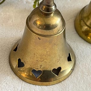 May include: Two brass bells with heart-shaped cutouts. The bells have a tarnished, vintage appearance. The bell on the left is in focus, showing the heart cutouts around the base. The bells are likely decorative items.