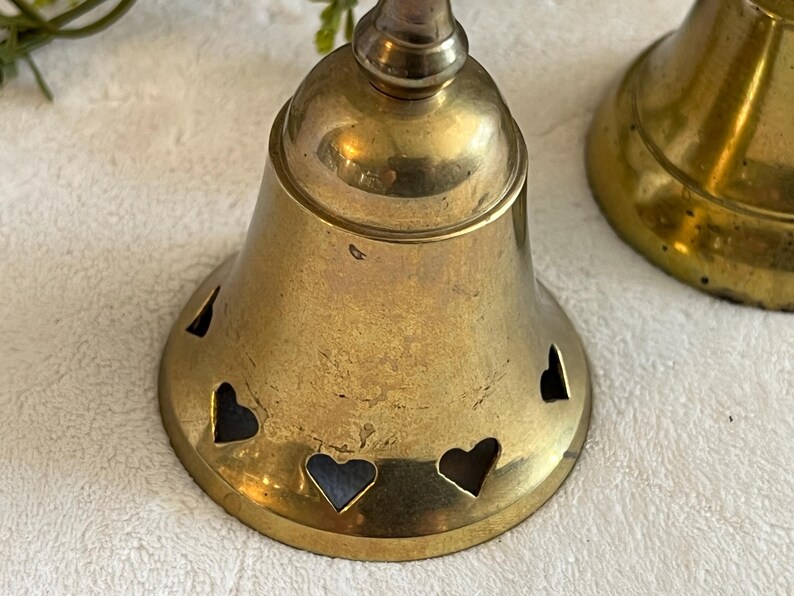 May include: Two brass bells with heart-shaped cutouts. The bells have a tarnished, vintage appearance. The bell on the left is in focus, showing the heart cutouts around the base. The bells are likely decorative items.