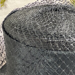 May include: A black hat with a rounded crown and a wide brim, covered with a fine, shimmering netting. The hat's texture appears woven, with a subtle sheen. The netting drapes over the hat, adding a layer of delicate detail.