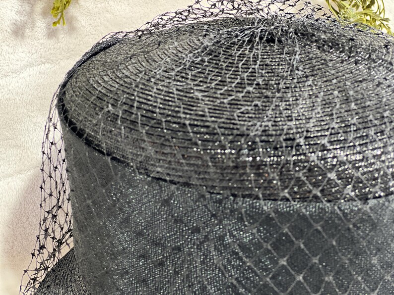 May include: A black hat with a rounded crown and a wide brim, covered with a fine, shimmering netting. The hat's texture appears woven, with a subtle sheen. The netting drapes over the hat, adding a layer of delicate detail.