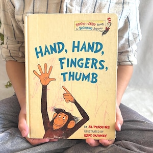 Può includere: Un libro per bambini vintage intitolato "Hand, Hand, Fingers, Thumb" con una copertina giallo chiaro. La copertina presenta un'illustrazione di scimmia dei cartoni animati e testo in blu e nero. Il libro fa parte della serie "Bright & Early Books for Beginning Beginners".