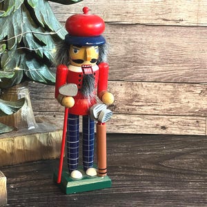 Könnte beinhalten: Eine hölzerne Nussknacker-Figur als Golfer verkleidet, mit rotem Barett, roter Jacke mit schwarzen Knöpfen und dunkelblau karierter Hose. Die Figur hat einen schwarzen Schnurrbart, graues Haar und hält einen Golfschläger auf einem grünen Sockel.