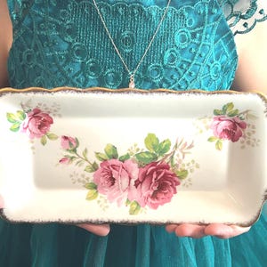 May include: A rectangular, off-white ceramic serving tray with a gold-trimmed, scalloped edge. The tray is decorated with pink roses and green leaves. The tray is being held by a person.