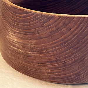 May include: A close-up of a large, round wooden bowl. The bowl is made of dark brown wood with visible grain patterns. The bowl's exterior has a smooth, curved surface.