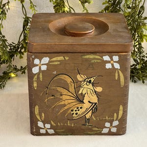 Puede incluir: Una caja de madera cuadrada con tapa, con un diseño de gallo pintado a mano en amarillo y marrón. Acentos florales blancos y detalles dorados adornan la caja. La tapa tiene una hendidura circular.