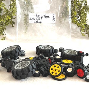 Op de afbeelding: Een verzameling zwarte Lego-banden in verschillende maten, sommige met gele en rode accenten. De afbeelding bevat een zak met het label "Leap Tires Sets of 4 6.9 oz". De banden liggen verspreid op een wit oppervlak.