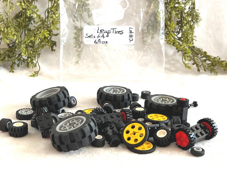 Op de afbeelding: Een verzameling zwarte Lego-banden in verschillende maten, sommige met gele en rode accenten. De afbeelding bevat een zak met het label "Leap Tires Sets of 4 6.9 oz". De banden liggen verspreid op een wit oppervlak.