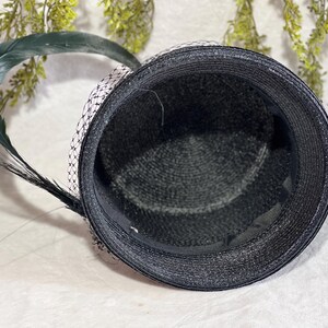 May include: Black hat with a textured weave, adorned with a dark green feather and a delicate black and white net veil. The hat's interior is lined with a smooth, dark fabric, creating a classic and elegant accessory.