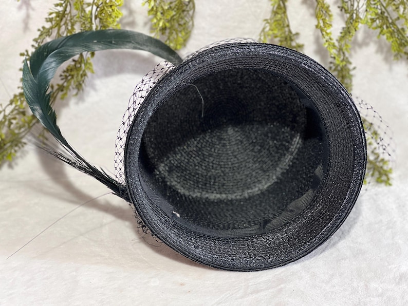 May include: Black hat with a textured weave, adorned with a dark green feather and a delicate black and white net veil. The hat's interior is lined with a smooth, dark fabric, creating a classic and elegant accessory.