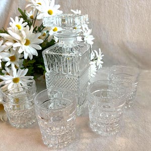 May include: A clear, cut-glass decanter with a stopper, accompanied by four matching lowball glasses. The set is arranged with a bouquet of white daisies, creating an elegant display. The decanter is square with a diamond pattern.