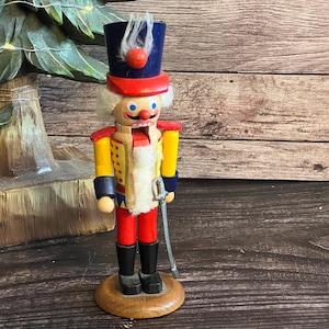 May include: A wooden nutcracker figurine featuring a blue hat with a white plume, a yellow jacket with red accents, red trousers, and black boots. The figure has a white beard, a painted face, and holds a silver-colored sword at its side.