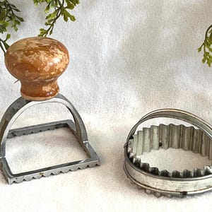 May include: Two vintage kitchen tools: a square cookie stamp with a wooden handle and a fluted metal biscuit cutter. The stamp has a wooden knob handle and a metal square base with a decorative edge. The cutter is a round metal band with a fluted edge.