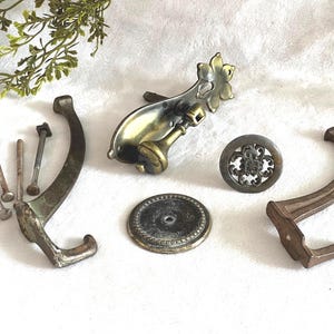 May include: An assortment of vintage metal hardware pieces, including hooks, knobs, and screws, in various shades of bronze and brass. The hardware is arranged on a white surface, with a sprig of greenery in the background.