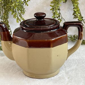 Puede incluir: Tetera de cerámica marrón de dos tonos con tapa. La tetera tiene una base de color beige claro y una sección superior y tapa de color marrón oscuro. El asa y el pico también son marrones. Un artículo de cocina vintage.