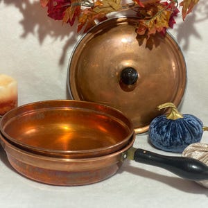 May include: A copper frying pan with a black handle and a copper lid. The pan is surrounded by decorative velvet pumpkins and candles, with fall foliage in the background.
