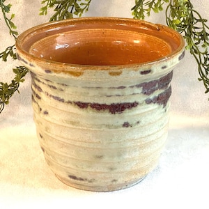 May include: A handmade ceramic planter with a wide, slightly flared rim. The exterior features a cream-colored base with horizontal ridges and irregular maroon and brown accents. The interior is a warm, earthy brown.