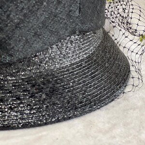 May include: Close-up of a black hat with a textured crown and a wide, shimmering brim. A delicate black netting veil drapes over the hat, adding a touch of elegance. The hat's design suggests a vintage or formal style.