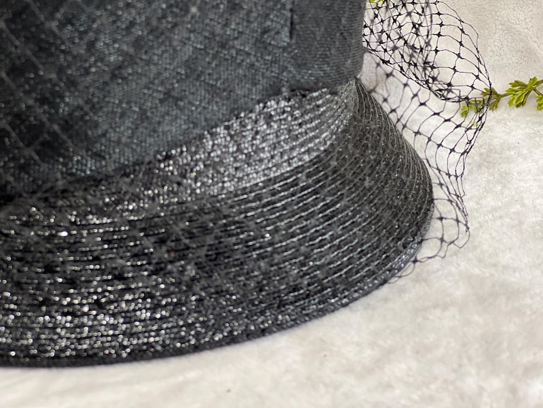 May include: Close-up of a black hat with a textured crown and a wide, shimmering brim. A delicate black netting veil drapes over the hat, adding a touch of elegance. The hat's design suggests a vintage or formal style.
