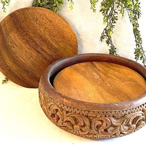Op de afbeelding: Een houten kom met een deksel, met ingewikkelde gesneden bloemmotieven. De kom heeft een warme bruine kleur, met een gladde, gepolijste afwerking. Het deksel is rond en ligt naast de kom.