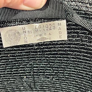 May include: Close-up of a black hat with a textured surface. A tag is sewn inside the hat, displaying the text "UNION MADE IN U.S.A." along with a serial number. The hat's brim is lined with a black ribbon.
