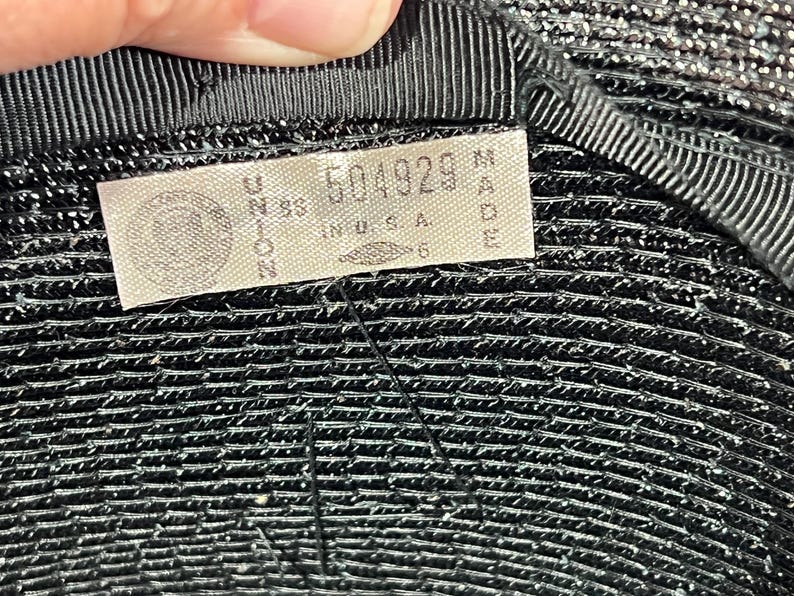 May include: Close-up of a black hat with a textured surface. A tag is sewn inside the hat, displaying the text "UNION MADE IN U.S.A." along with a serial number. The hat's brim is lined with a black ribbon.