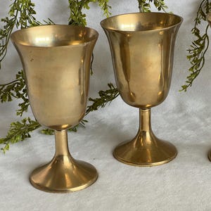 May include: Three vintage gold-colored goblets with a tulip-shaped bowl and a pedestal base. The goblets are made of metal and have a shiny finish. The background includes greenery.