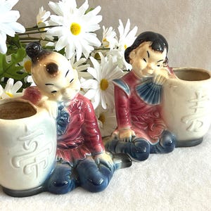 May include: Two ceramic planters shaped like figures kneeling beside cylindrical vessels. The figures wear red and blue clothing, and the cream-colored vessels feature embossed characters. The planters have a glossy finish and painted details.