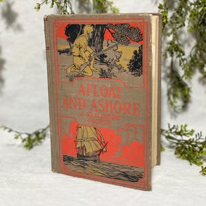 Può includere: Libro d'epoca intitolato "Afloat and Ashore" di J. Fenimore Cooper. La copertina beige presenta illustrazioni arancioni e gialle, tra cui un uomo con un fucile e una nave a vela. Il titolo è in lettere rosse.