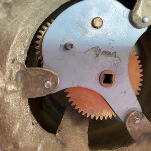 Op de afbeelding: Close-up van de binnenkant van een vintage klok. Het mechanisme heeft messing tandwielen en een zilverkleurige plaat met een vierkant gat in het midden.