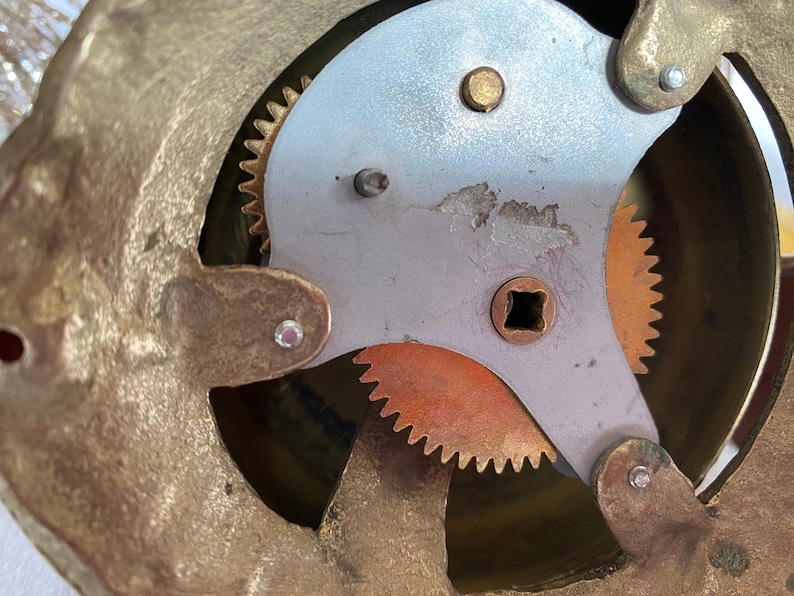 Op de afbeelding: Close-up van de binnenkant van een vintage klok. Het mechanisme heeft messing tandwielen en een zilverkleurige plaat met een vierkant gat in het midden.