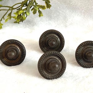 May include: Four antique bronze-colored cabinet knobs with a circular design. Each knob features a central rounded element surrounded by concentric circles and a beaded outer edge. The knobs are arranged on a white surface with green foliage in the background.